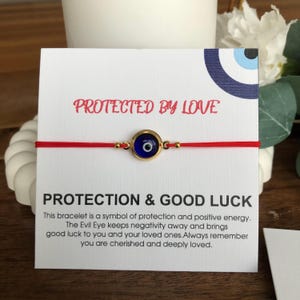 Puede incluir: Pulsera de hilo rojo con un dije de ojo turco dorado. El dije tiene un centro azul con una pupila negra. La tarjeta detrás de la pulsera dice "PROTECTED BY LOVE" y "PROTECTION & GOOD LUCK".