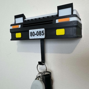 May include: A black and white key holder shaped like a car front, with a license plate that reads "80-085". The key holder has yellow and orange accents, and a black hook for keys. The key holder is mounted on a white wall.