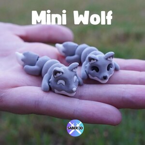 May include: Two gray, miniature wolf figurines with white-tipped tails and black eyes are displayed. The text "Mini Wolf" is above the figurines. The figurines are resting on a person's palm, with a blurred green background.