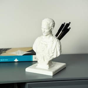 May include: White bust sculpture of a man wearing a toga, used as a pen holder. The bust is on a white base and has three black pens sticking out of the back of the head.
