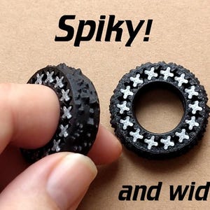 May include: Two black, spiky, and wider rings with white cross-shaped accents. The text "Spiky!" and "and wider" are displayed above and below the rings, respectively. The rings are on a tan surface.