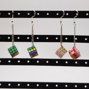 May include: Four pairs of dangle earrings featuring miniature Rubik's Cubes. Each cube is a different color combination, suspended from silver-tone chain and hook ear wires. The earrings are displayed against a black and white background.