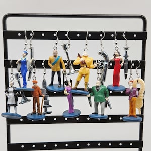 May include: A set of 10 pairs of dangle earrings featuring miniature figurines. The figurines are dressed in various outfits and hold different accessories. The earrings are displayed on a black earring stand.