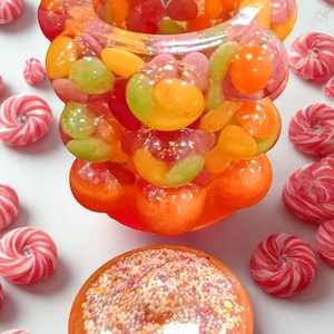 Peut inclure: Une sculpture en résine orange remplie de bonbons gélifiés colorés et une base en forme de donut. La sculpture est entourée de bonbons tourbillonnants rouges et blancs, créant une esthétique ludique.