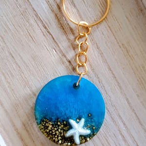 May include: A round keychain with a blue resin design depicting an ocean scene. The design includes a white starfish and gold-colored sand. The keychain has a gold-colored ring and chain.