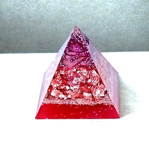 Puede incluir: Un objeto decorativo en forma de pirámide con un degradado de colores del rojo al rosa. La pirámide está rellena de papel de aluminio rojo y purpurina, y la base es de un rojo intenso. El objeto está sobre una superficie blanca.