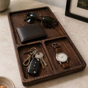 May include: A rectangular dark brown wooden valet tray with three divided compartments. The tray holds a pair of dark-rimmed sunglasses, a dark brown leather wallet, a set of metal keys with a black key fob, and a wristwatch with a brown leather strap.