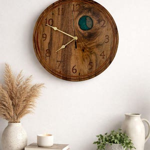 May include: A round wooden wall clock with a natural wood grain finish. The clock features gold-colored hands and a teal-colored accent. The clock is surrounded by decorative items, including a vase with dried plants.
