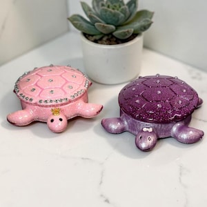 May include: Two turtle-shaped trinket boxes, one in light pink and one in deep purple, both featuring a glittery finish and embedded clear rhinestones on their shells. The pink turtle has a small gold crown on its head, while the purple turtle has a small white bow on its head.