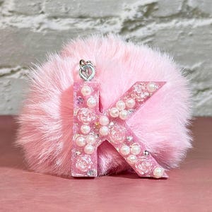 May include: A pink resin letter "K" keychain with embedded pearls and floral accents, attached to a fluffy pink pom-pom. The keychain has a silver-colored clasp.
