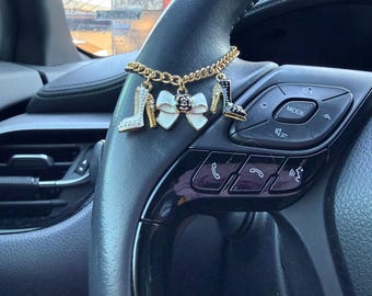 May include: A gold-toned metal chain charm accessory featuring three dangling charms: a white high-heeled shoe, a white bow with a central black flower detail, and a black high-heeled shoe, all attached to a steering wheel.