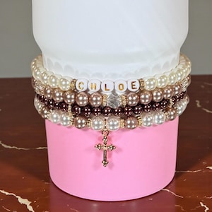 May include: A stack of beaded bracelets in shades of white, brown, and gold, with the name "CHLOE" spelled out in gold letters. A gold cross charm hangs from the bottom bracelet. The bracelets are displayed on a pink surface.