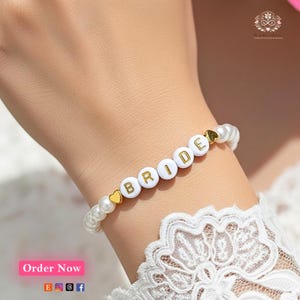 Könnte beinhalten: Ein weißes Perlenarmband mit der Aufschrift "BRIDE" und goldenen Herzakzenten. Das Armband besteht aus weißen Perlen und goldenen Buchstabenperlen. Das Armband ist ein Brautaccessoire.