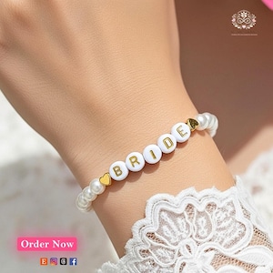May include: A beaded bracelet featuring white round beads and gold-colored letter beads that spell out the word BRIDE. The bracelet includes two small gold-colored heart-shaped beads placed at the ends of the letter sequence.