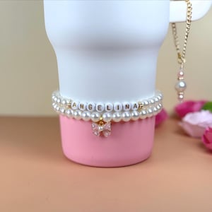 Puede incluir: Un vaso blanco con base rosa está adornado con una pulsera de perlas de doble hebra que deletrea "ANGELINA" en cuentas de letras. Un pequeño dije de lazo cuelga debajo. También se ve un collar de cadena dorada con un colgante de perla.