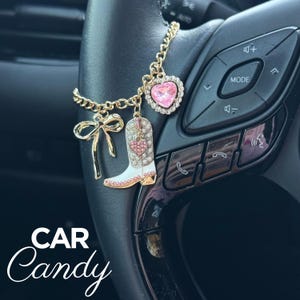 May include: Gold-toned car charm featuring a pink heart-shaped pendant, a white cowboy boot with a pink heart, and a bow. The charm hangs from a steering wheel. The text "CAR Candy" is at the bottom.