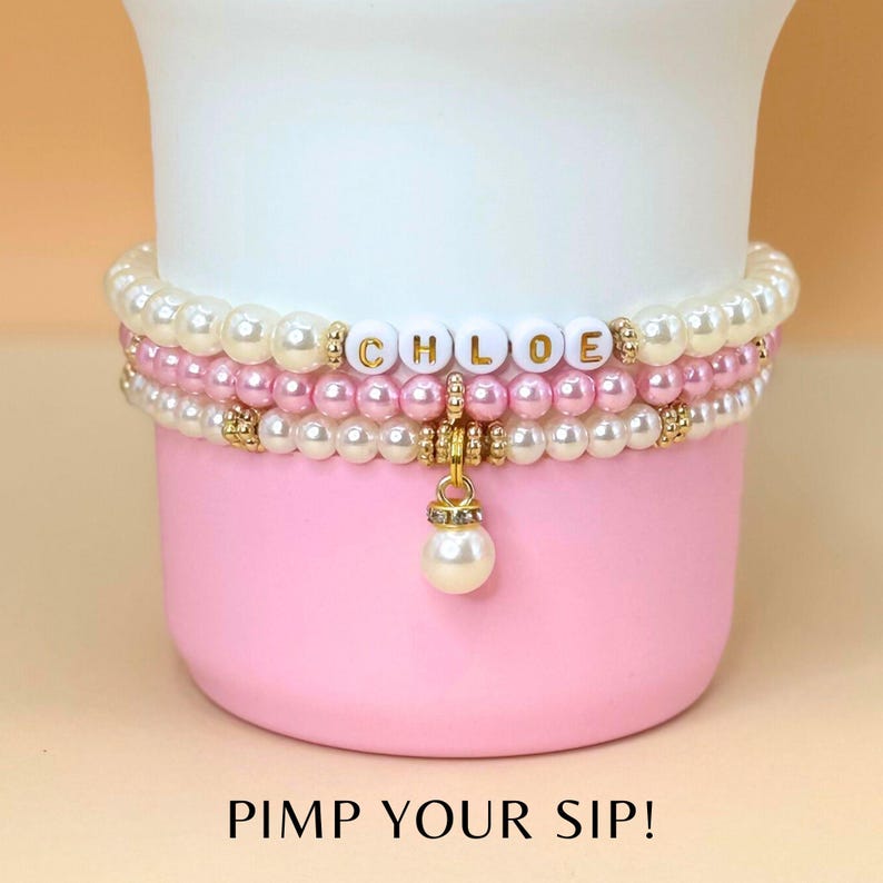 May include: A white cup with a pink rim and the text "PIMP YOUR SIP!" printed on the bottom. The cup is decorated with a three-strand pearl bracelet with a gold charm and the name "CHLOE" spelled out in white letters.