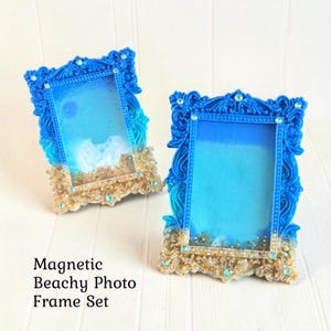 Op de afbeelding: Een set van twee rechthoekige magnetische fotolijsten met sierlijke blauwe harsranden, ingewikkeld krulwerk en ingebedde lichtblauwe strass-steentjes. De onderste delen bevatten zandachtig materiaal. Tekst: Magnetic Beachy Photo Frame Set.