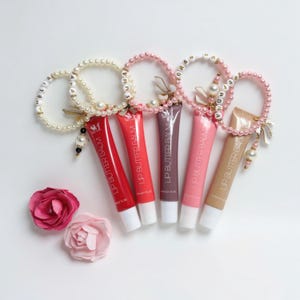 Könnte beinhalten: Eine Auswahl an Lippenbutter-Balsam-Tuben in verschiedenen Rot-, Rosa- und Brauntönen, die jeweils an einem Perlenarmband befestigt sind. Die Tuben sind mit zwei Stoffrosen auf einer weißen Oberfläche angeordnet. Der Text "LIP BUTTER BALM" ist auf den Tuben zu sehen.