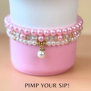 May include: A pink cup with a white top, adorned with three beaded bracelets. The top bracelet features pink pearls, the middle one has clear beads and gold accents, and the bottom one has white pearls. A pearl charm hangs from the bottom bracelet. The text "PIMP YOUR SIP!" is at the bottom.