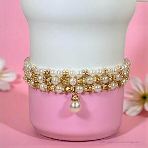 May include: A white and pink tumbler with three rows of faux pearl and gold beaded bracelets. A single pearl charm hangs from the bottom bracelet. The background is pink with white flowers.
