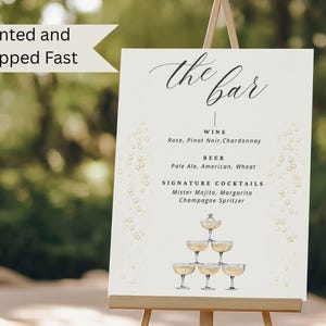 May include: A cream-colored sign on an easel with the words "the bar" in elegant script. The sign lists wine, beer, and signature cocktails, including Mister Mojito and Margarita. Gold bubbles and champagne glasses decorate the sign.