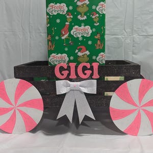 May include: A black wooden wagon with pink and white peppermint candy wheels. The wagon is decorated with glitter and the name "GIGI" in pink letters. The wagon is decorated with wrapping paper featuring a green Grinch character.