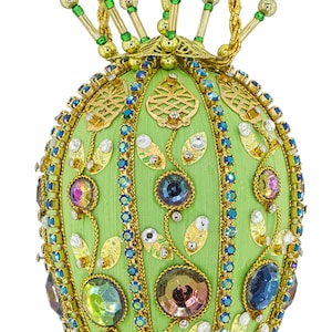May include: An ornate, egg-shaped ornament with a light green fabric base. It is embellished with gold-colored metal filigree, colorful gemstones, and blue and green beads. A gold chain is attached for hanging.