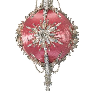 May include: A pink, orb-shaped Christmas ornament with a textured surface. The ornament is embellished with silver-colored metal, clear crystals, and faux pearls. A starburst design is centered on the front.