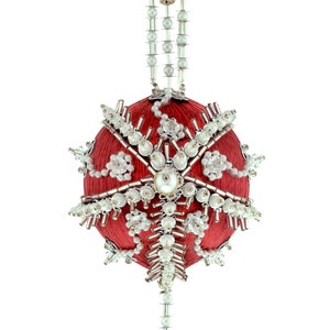 May include: A red Christmas ornament with intricate beadwork. The ornament is round, with a central pearl and crystal accents. It hangs from a beaded silver chain, creating a festive and elegant holiday decoration.