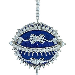 May include: A blue, oval-shaped ornament with intricate detailing. The ornament features a dark blue fabric base, embellished with white beads, silver accents, and crystal-like pendants. Decorative bows are also present.