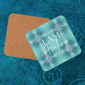 Op de afbeelding: Vierkante onderzetter met een turquoise achtergrond en witte sneeuwvlokachtige patronen. De woorden "I AM Healing" zijn in witte letters geschreven. Ernaast ligt een bruine kurken onderzetter.