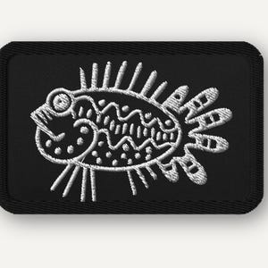 May include: A rectangular black embroidered patch featuring a white stitched line art design of a stylized fish. The fish has a round eye, a wavy body pattern, and spiky fins. The patch has a thick black stitched border with rounded corners.