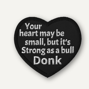Puede incluir: Parche bordado negro en forma de corazón con texto blanco: "Your heart may be small, but it's strong as a bull Donk." El parche tiene un borde negro y es probablemente para ropa o accesorios.