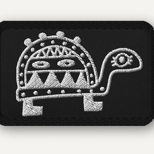 May include: A rectangular black embroidered patch featuring a white stitched outline of a stylised turtle. The turtle shell displays geometric patterns including triangles and dots, while the head features a single eye with eyelashes.
