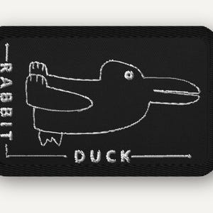 Pode incluir: Um emblema retangular preto bordado com uma ilustração de linhas brancas da clássica ilusão de ótica coelho-pato. A palavra RABBIT está bordada verticalmente no lado esquerdo e a palavra DUCK está bordada horizontalmente na parte inferior.