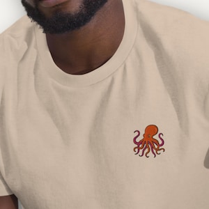 May include: A beige t-shirt with a small embroidered octopus design in orange, red, and purple. The octopus is on the chest. The shirt has a crew neck and short sleeves. The fabric appears to be cotton.