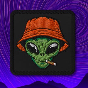 Op de afbeelding: Geborduurde patch met een groene alien met een oranje vissershoed en een sigaar. De alien heeft grote zwarte ogen en een serieuze uitdrukking. De patch is vierkant met een zwarte rand.