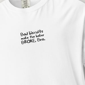 May include: White t-shirt featuring the phrase "Bad biscuits make the baker BROKE, Bro." in black. The shirt has a crew neck and short sleeves. The tag reads "COMFORT COLORS XL-TG-2X".