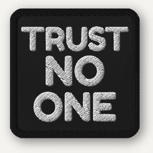 Może przedstawiać: Czarna kwadratowa naszywka z wyhaftowanym białym napisem "TRUST NO ONE". Naszywka ma czarną obwódkę i teksturowany wygląd, co sugeruje, że jest wykonana z tkaniny.