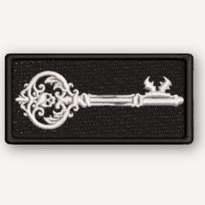 May include: A rectangular embroidered patch featuring a white skeleton key design on a black background. The key has an ornate, filigree-style bow with a small skull detail at the centre and a jagged bit at the end, all outlined in black.