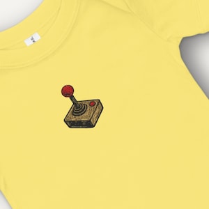 May include: Yellow baby bodysuit with a retro video game joystick graphic. The joystick has a red ball on top and is set on a brown square base. The bodysuit is made of soft material.