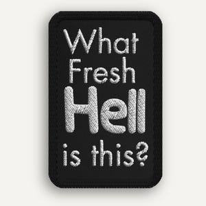 May include: Black rectangular embroidered patch with the phrase "What Fresh Hell is this?" in white thread. The patch has a black border and is likely designed to be sewn or ironed onto fabric.