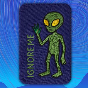 May include: Embroidered patch featuring a green alien with large yellow eyes and a waving hand. The alien is on a navy blue background with the words "IGNORE ME" in green. The patch has a rectangular shape with rounded corners.