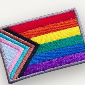 May include: A rectangular embroidered patch featuring the Progress Pride flag design. The patch displays horizontal stripes of red, orange, yellow, green, blue, and purple, with a chevron on the left side in black, brown, light blue, pink, and white.