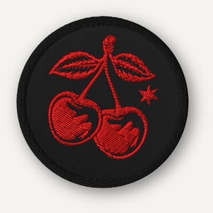 Puede incluir: Un parche redondo bordado en negro con un par de cerezas rojas, hojas verdes y una estrella. Las cerezas están detalladas con hilo rojo, y el parche tiene un borde negro.