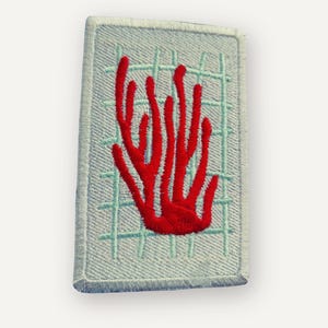 May include: A rectangular embroidered patch featuring a bright red coral-like branching design centered on a light blue background with a grid pattern of thin, pale green lines. The patch has a stitched border and a textured fabric surface.