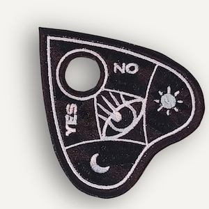 May include: A black heart-shaped embroidered patch featuring white stitched details including a central eye, a sun, a crescent moon, and the words YES and NO. The patch has a circular cutout near the top and a white border outlining the entire shape.