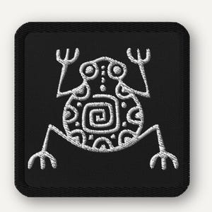 May include: Square black patch with a white embroidered frog design. The frog has a stylized, symmetrical appearance with a spiral on its body. The patch has rounded corners and a black border.