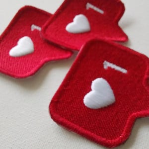 May include: Three red fabric patches with a white heart and a white number 1 embroidered on each. The patches have a unique shape with a pointed bottom and a curved top. The patches are on a white surface.